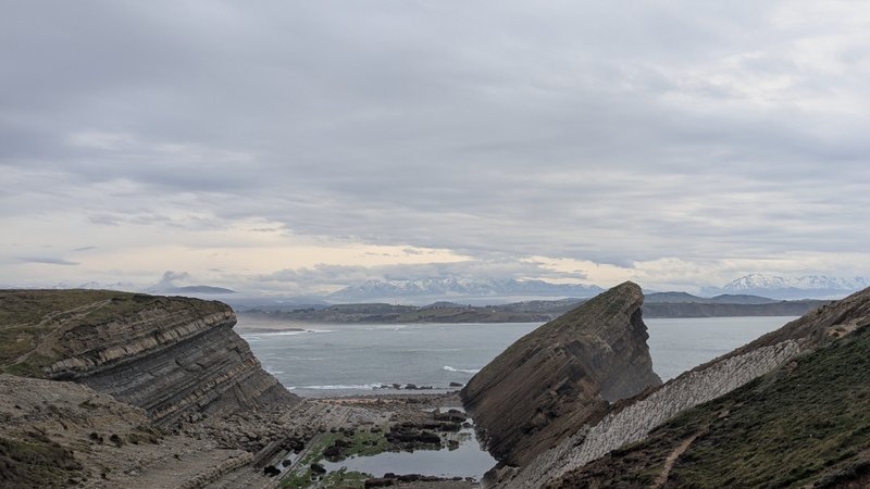 A wide view of Mogro Bay showing distinctive rock formations and distant snowy mountains under a cloudy sky. — Ria de Mogro (Baía de Mogro), Mogro, Cantabria, Spain