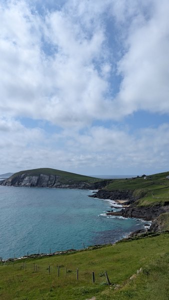 A scenic view of a bay with turquoise water, rocky shorelines, green hills, and a small sandy beach under a cloudy sky. — Coumeenole (Dunquin), Dunquin, Kenmare Municipal District, Ireland