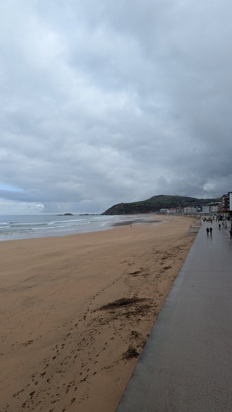 A wide view of Zarautz beach and promenade on an overcast day, with small waves breaking.