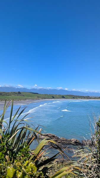Tauranga Bay features a wide sandy beach with waves, backed by green hills, under a clear blue sky. — Baía de Tauranga (Westport), Tauranga Bay, West Coast, New Zealand
