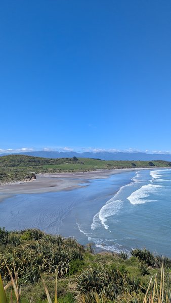An aerial view of Tauranga Bay showing the curving beach and ocean waves under a clear sky. — Baía de Tauranga (Westport), Tauranga Bay, West Coast, New Zealand