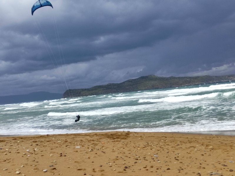 A kitesurfer riding waves at Stalos beach under a cloudy sky.