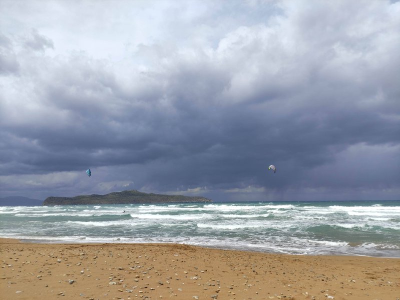 Kitesurfers riding in wavy conditions at Stalos beach under a cloudy sky.