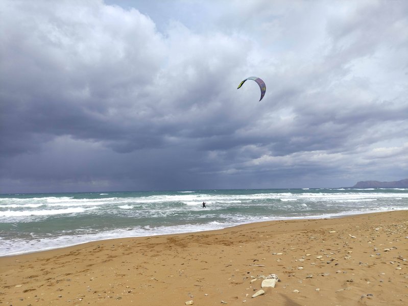 A kitesurfer rides in wavy conditions at Stalos beach under a stormy sky.