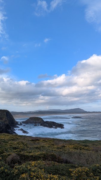 A wide view of the Valdoviño coastline showing rocky outcrops, breaking waves, and a distant beach under a partly cloudy sky. — Valdoviño (Da Frouxeira), Valdoviño, Galicia, Spain