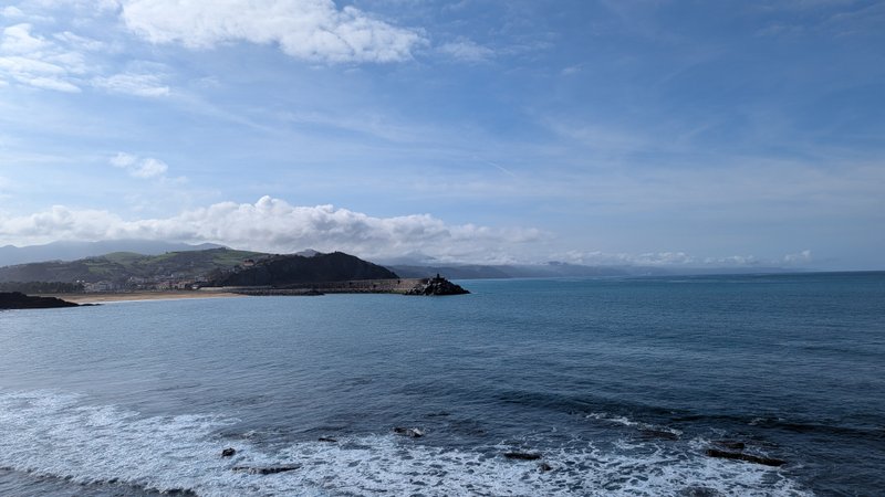 A panoramic view of the coastline showing the bay, beach, and distant town with mountains under a partly cloudy sky. — Orrua (Getaria), Getaria, Autonomous Community of the Basque Country, Spain