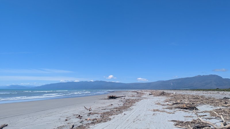 A wide sandy beach covered in driftwood meets the ocean with mountains visible in the distance under a clear blue sky. — Shingles (Westport), Westport, West Coast, New Zealand
