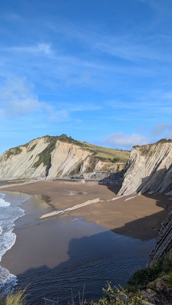 A wide view of the sandy beach and the distinct stratified cliffs of Zumaya, with calm waves rolling in.