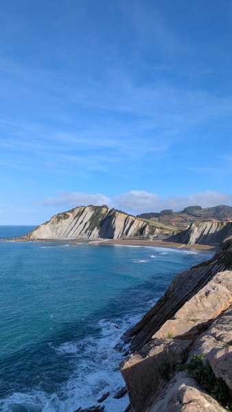 A scenic view of the stratified cliffs, sandy beach, and clear blue waters of Zumaya Bay.