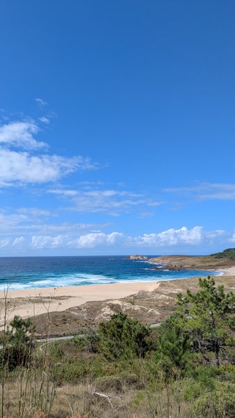 A wide view of Playa de Doninos showing the sandy beach, ocean with waves, and a distant rocky coastline under a blue sky.