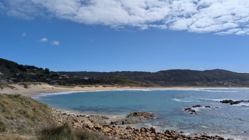 A panoramic view of Playa de Doninos with a sandy beach, clear blue water, and rocky outcrops under a blue sky.