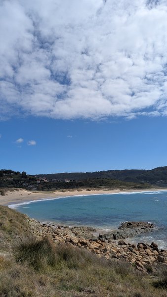 An elevated view of the sandy beach and rocky coastline of Playa de Doninos under a partly cloudy sky.