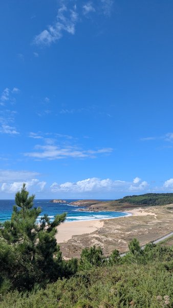 An expansive view of Playa de Doninos beach with sandy dunes, breaking waves, and distant headlands under a blue sky.
