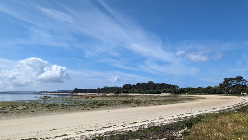 A wide sandy beach at A Illa de Arousa with shallow water and a treeline backdrop.