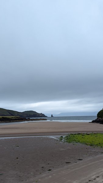 A wide view of Inch Reef beach with a sandy shore, distant hills, and a prominent rock formation in the sea under an overcast sky.