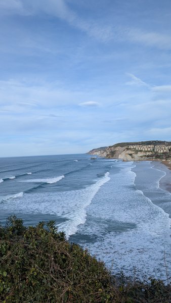 An aerial view shows waves rolling onto a long sandy beach bordered by cliffs with buildings in the distance. — La Salvaje, Sopela, Autonomous Community of the Basque Country, Spain