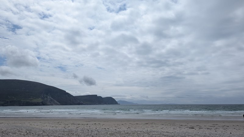 A wide view of Keel beach and the ocean, backed by green hills under a cloudy sky. — Keel (Ilha de Achill), County Mayo, Westport-Belmullet Municipal District, Ireland