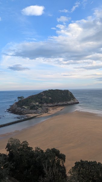 An aerial view shows a sandy beach, a river mouth, and an island covered in trees, meeting the ocean under a cloudy sky. — Praia de Carraspio (Mendexa), Mendexa, Autonomous Community of the Basque Country, Spain