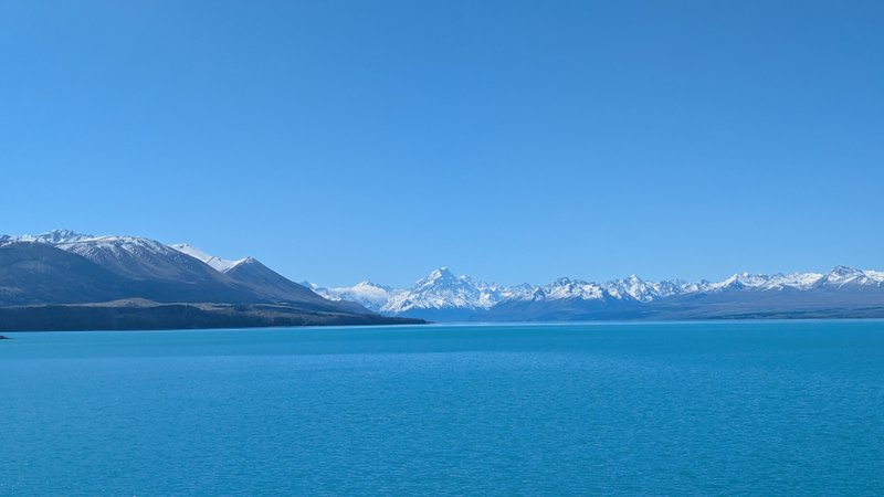 A wide view of Lake Pukaki with snow-capped mountains under a clear blue sky. — Ponto Lake Pukaki (Lago Pukaki), Mackenzie District, Canterbury, New Zealand