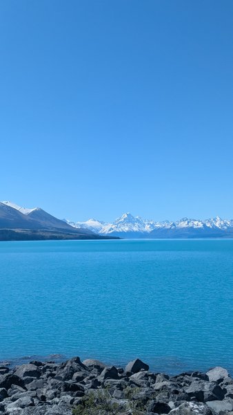 A wide shot of Lake Pukaki with its turquoise water, rocky shoreline, and snow-capped mountains in the background. — Ponto Lake Pukaki (Lago Pukaki), Mackenzie District, Canterbury, New Zealand