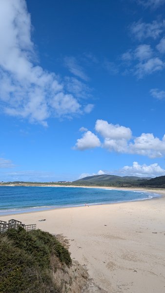 A wide view of a sandy beach curving around a bay with blue water and distant hills under a partly cloudy sky. — San Xurxo da Marina (Ferrol), O Porto, Galicia, Spain
