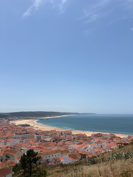 An elevated panoramic view of Praia da Nazaré, showing the town, wide sandy beach, and the Atlantic Ocean.