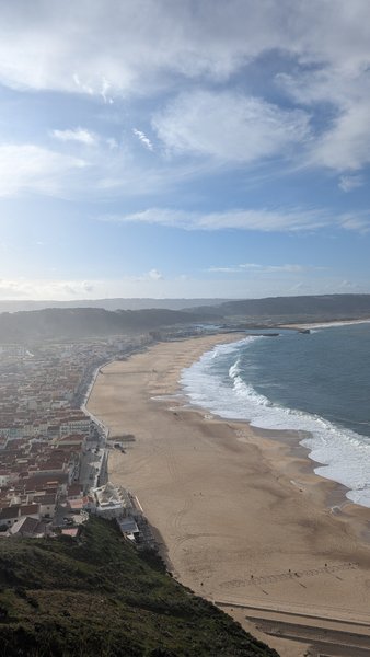 An aerial view shows the long sandy beach of Nazaré with waves breaking and the town alongside. — Praia da Nazaré, Nazaré, Portugal