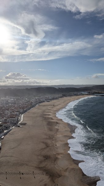 An expansive aerial view of Praia da Nazaré, showing the long sandy beach, breaking waves, and the town stretching along the coast.