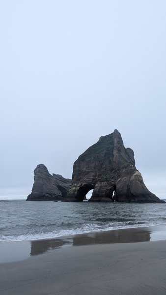 A large rock formation with a natural arch sits just offshore from a sandy beach under an overcast sky. — Praia de Wairaiki, Tasman District, Tasman, New Zealand