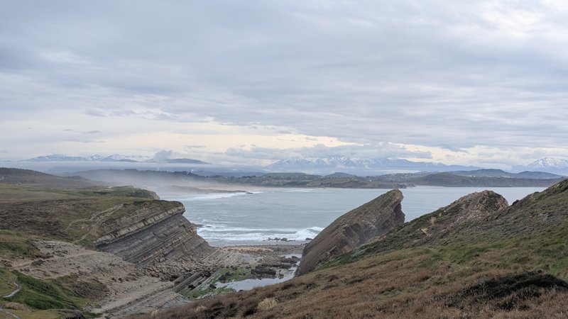 A wide panoramic view of the rocky coastline, beach, and bay under an overcast sky, with distant snow-capped mountains. — Liencres (Cantábria), Piélagos, Cantabria, Spain