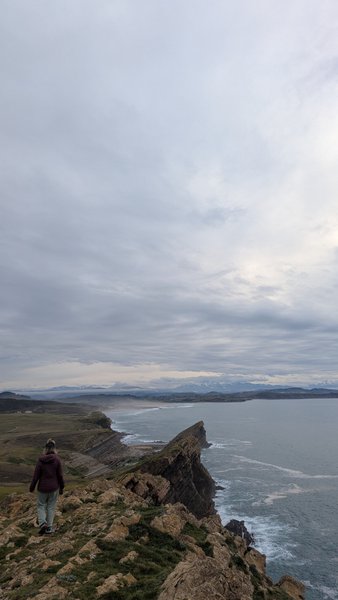 A person stands on a rocky clifftop overlooking the ocean and a distant sandy beach under a cloudy sky. — Liencres (Cantábria), Piélagos, Cantabria, Spain