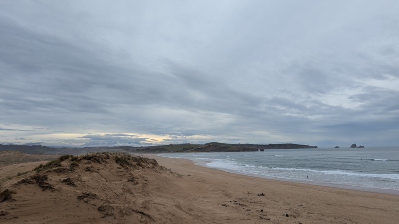 An expansive sandy beach with dunes and waves, looking towards distant cliffs and sea stacks under an overcast sky. — Liencres (Cantábria), Piélagos, Cantabria, Spain