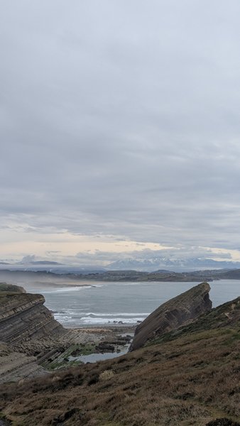A wide view of the rugged Liencres coastline with waves breaking in the bay under an overcast sky, with distant snowy mountains. — Liencres (Cantábria), Piélagos, Cantabria, Spain