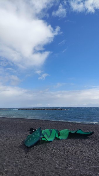 A green kite lies on a dark sand beach with the ocean and a breakwater in the distance. — Puerto de Güímar (Tenerife), Güímar, Canary Islands, Spain
