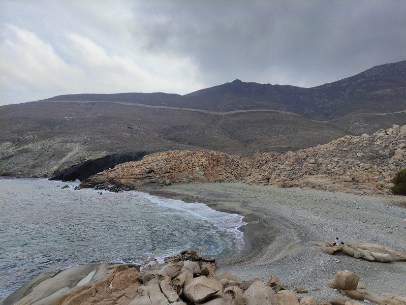 A wide view of Leivada beach showing the water, pebble shore, and rocky landscape under an overcast sky. — Leivada (Tinos), Tinos Regional Unit, South Aegean, Greece