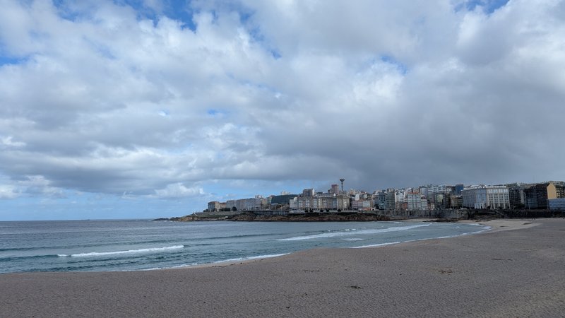 A wide view of Playa de Riazor showing the sandy beach, breaking waves, and the city skyline.