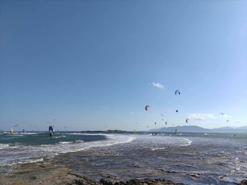 Numerous kitesurfers and windsurfers on the water at Nissakia Loutsa under a clear blue sky.