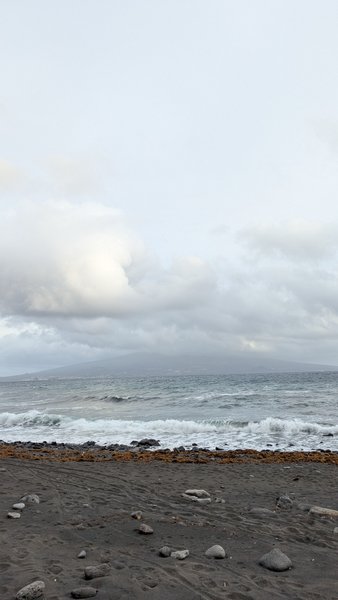A cloudy view of the dark sand beach and ocean with a distant landmass under an overcast sky. — Praia do Almoxarife (Almoxarife), Praia do Almoxarife, Portugal