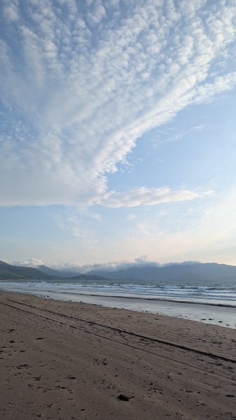 A wide sandy beach with gentle waves and mountains in the background under a textured sky. — Stoney Gap, Castlegregory, Kenmare Municipal District, Ireland