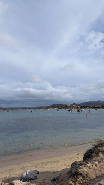 A wide view of the riding area at Isola Gabbiani with several windsurfers and wing foilers on the water. — Isola Gabbiani (Palau Coast), Lu Palau/Palau, Sardinia, Italy