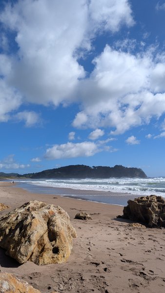 A wide view of Hot Water Beach showing breaking waves and the distant headland under a cloudy sky. — Praia de Água Quente, Hot Water Beach, Waikato, New Zealand