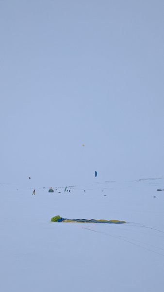 Snowkiters are visible on a vast, snow-covered plateau under a pale sky, with a packed kite in the foreground. — Batstjorn (Planície do Hardangervidda), Norway