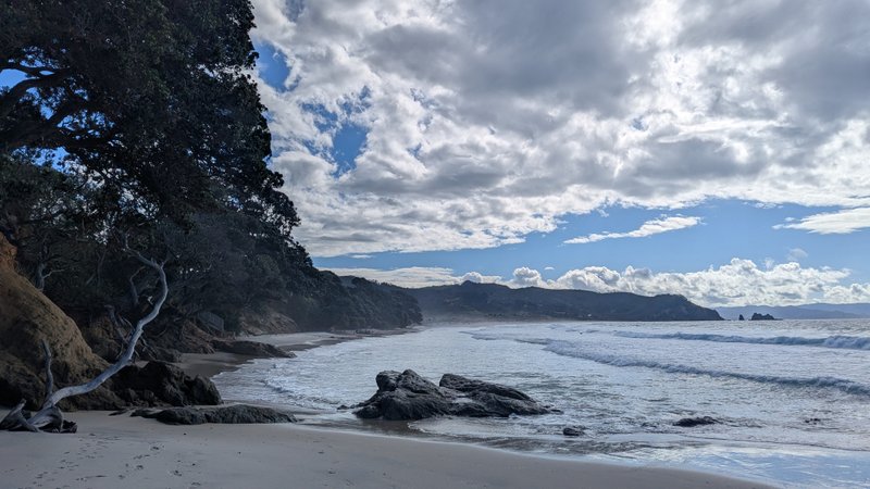A scenic view of Otama Beach with waves rolling onto the sand and a tree-lined coast under a partly cloudy sky.
