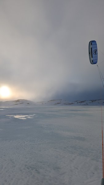 A kite flies over a vast, snow-covered winter landscape under a bright, cloudy sky with the sun low on the horizon. — Oerteren, Buskerud, Norway