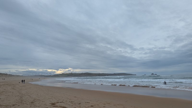 A wide sandy beach and ocean with a distant surfer under an overcast sky. — Playa de Robayera (Mogro Bay), Mogro, Cantabria, Spain